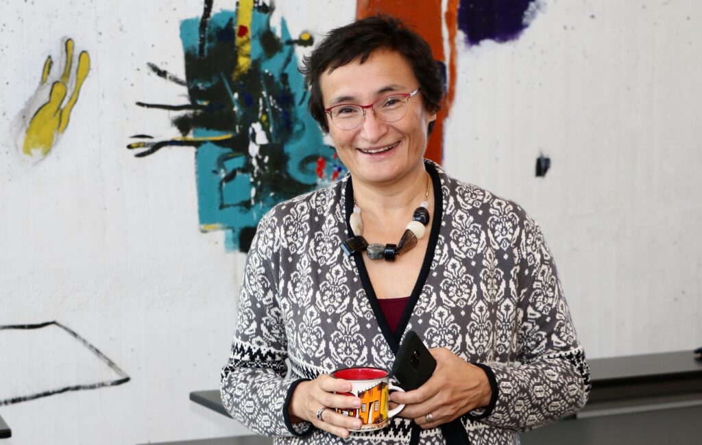 Virginia Dignum stands in front of a painted wall with a cup of coffee and a cell phone.