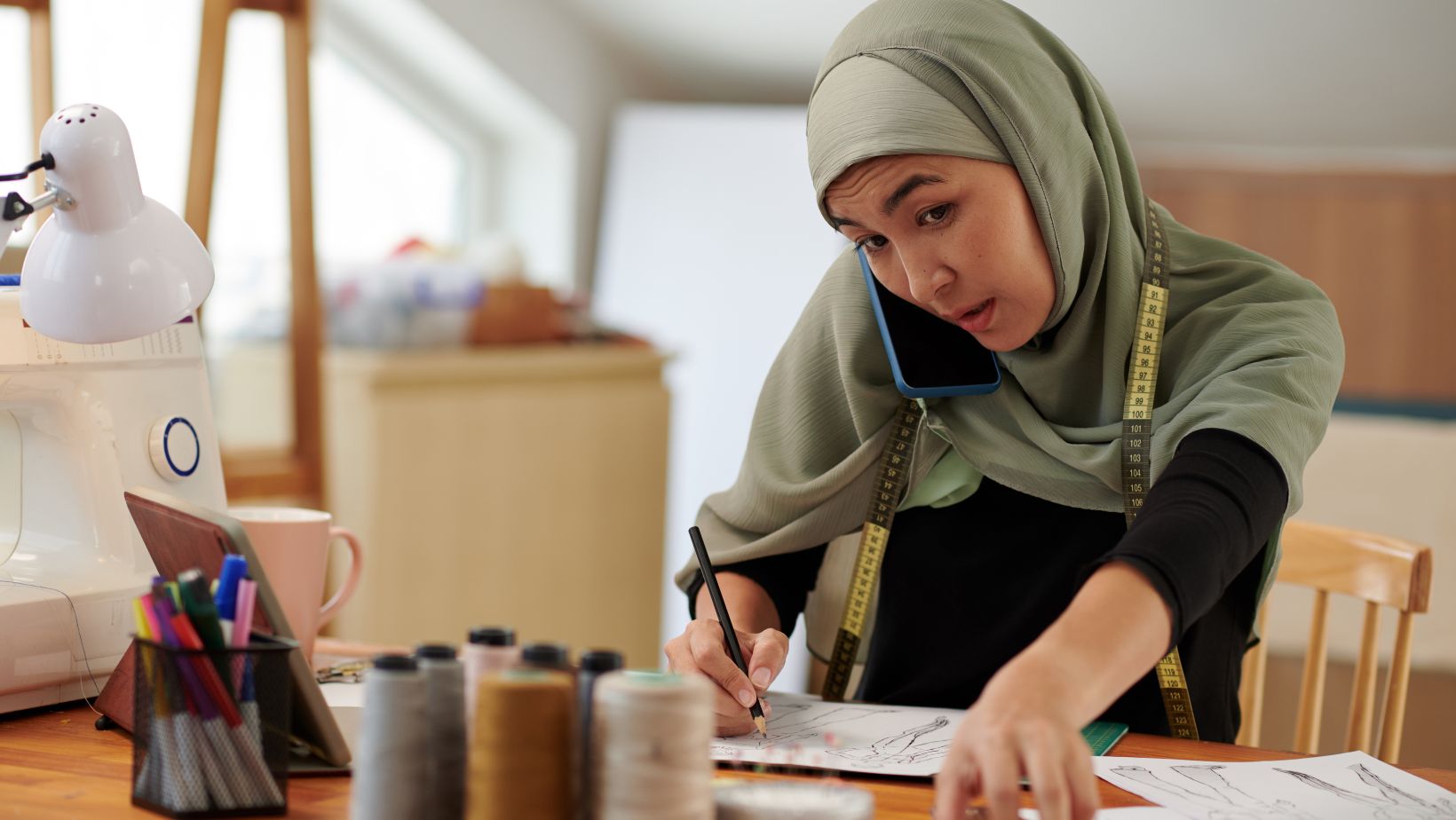 En kvinna (entreprenör) pratar i telefon samtidigt som hon skissar ett klädesplagg.